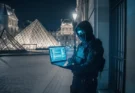 A masked man holding a laptop near the Louvre service entrance at night, symbolizing the cyberattack in the 2025 jewelry heist.